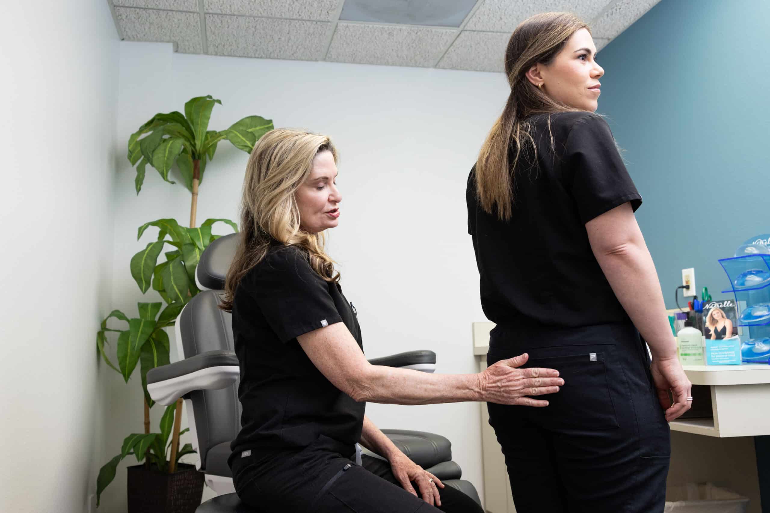 A healthcare professional examines a standing patient's lower back in a medical office. The professional is seated and gently touching the patient's lower back while providing guidance. The room features a potted plant, medical chair, and various medical supplies in the background.