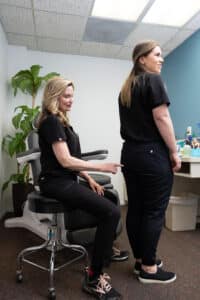 Two women are in an office setting. One woman is sitting on a stool, wearing black clothing, pointing at the other woman's lower back. The other woman, also dressed in black, is standing and facing away. There are office furniture and plants in the background. The room has a light-colored wall and a blue accent wall.