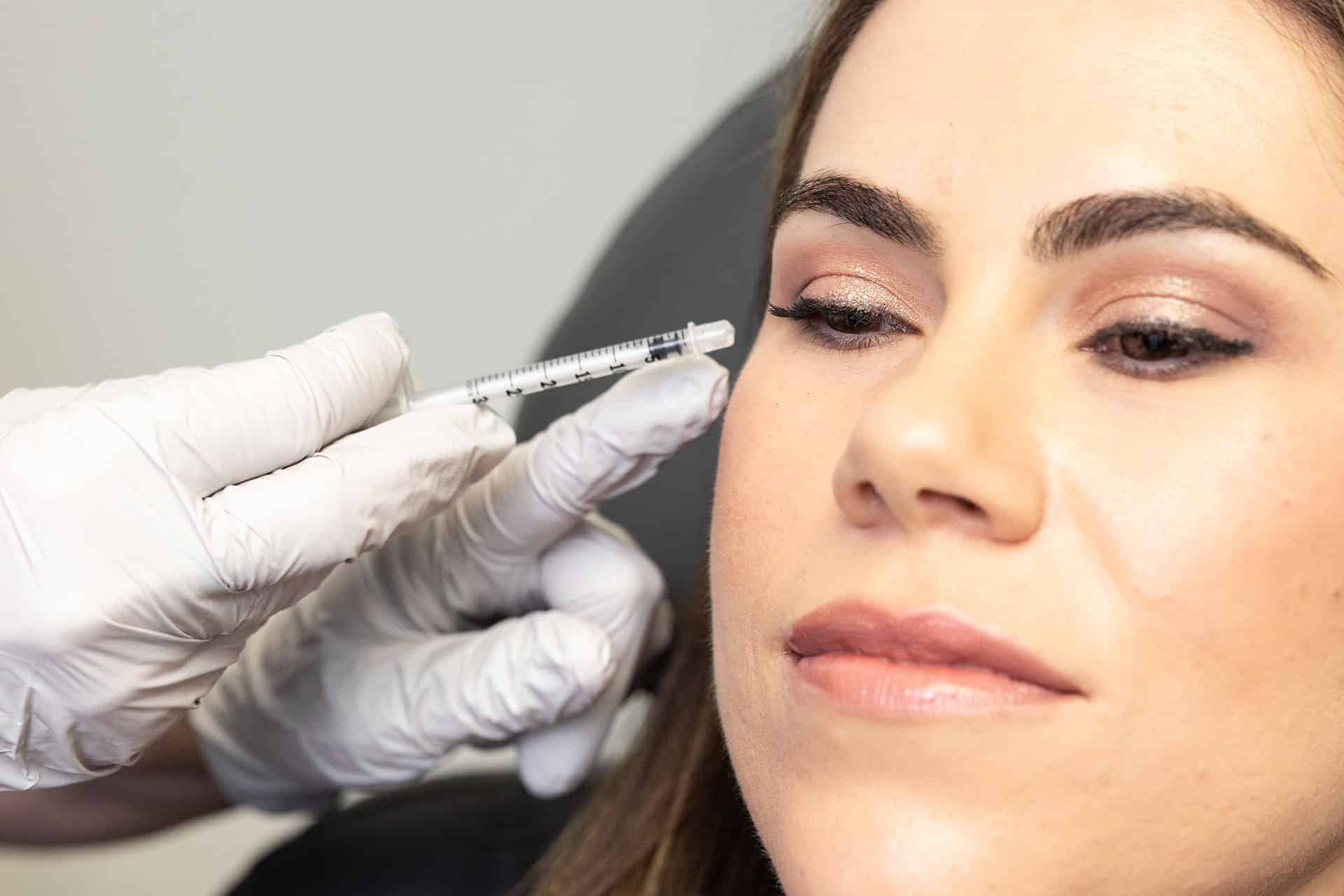 A close-up of a woman receiving a cosmetic injection on the side of her face near her eye. The person administering the injection is wearing white medical gloves and holding a syringe. The woman has her eyes closed and appears relaxed. The background is neutral and out of focus.