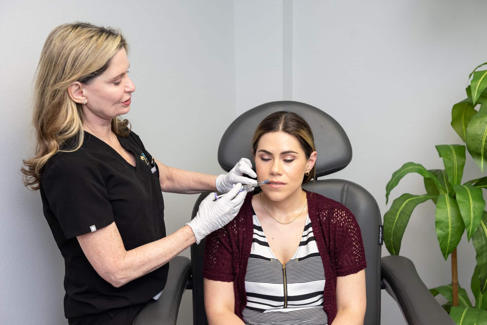 A healthcare professional wearing black scrubs and white gloves is administering an injection to a seated woman's cheek. The woman, who is wearing a striped top with a red cardigan, appears calm with her eyes closed. There is a potted plant in the background.