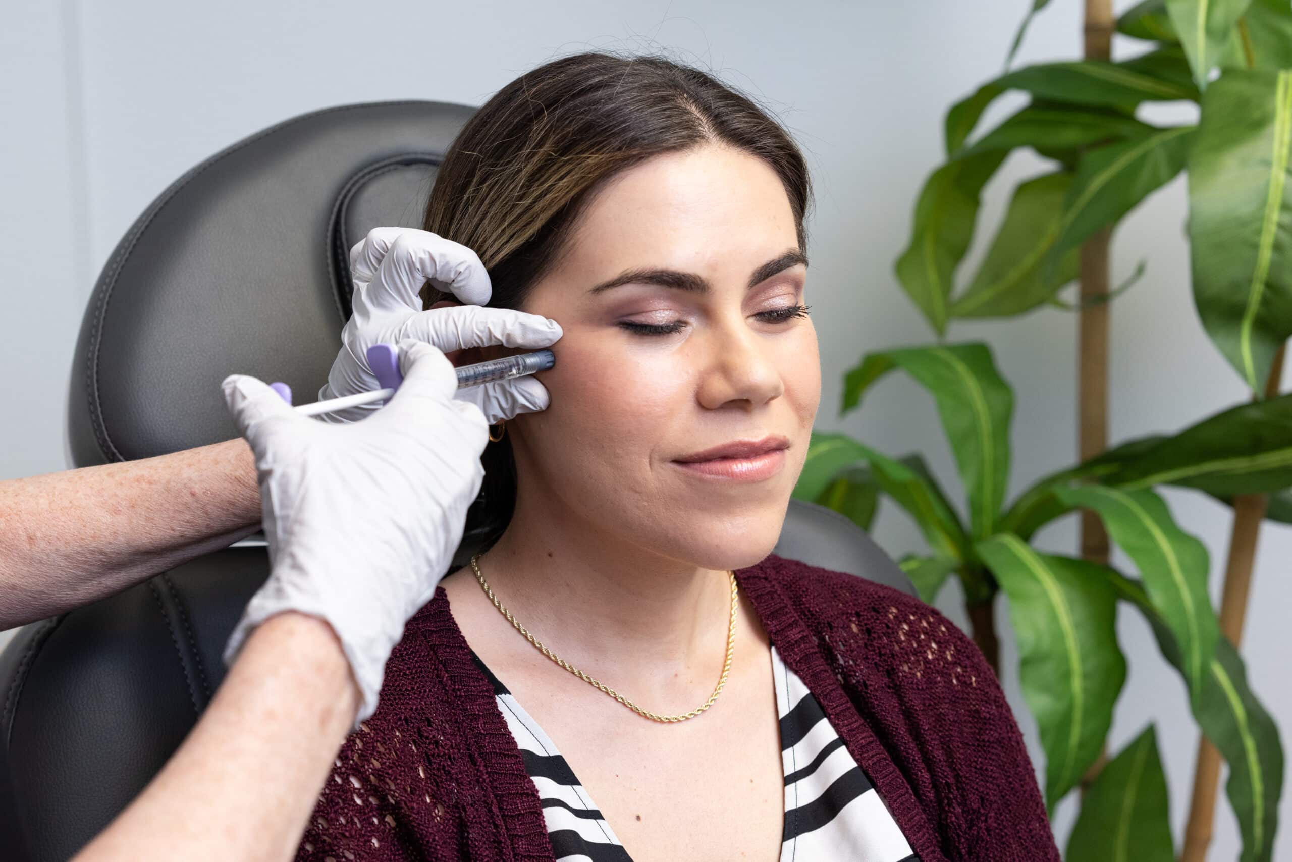 A person with closed eyes sits comfortably in a chair wearing a burgundy cardigan over a black and white striped shirt. A gloved hand is administering a facial injection to their cheek. There is a green plant in the background.