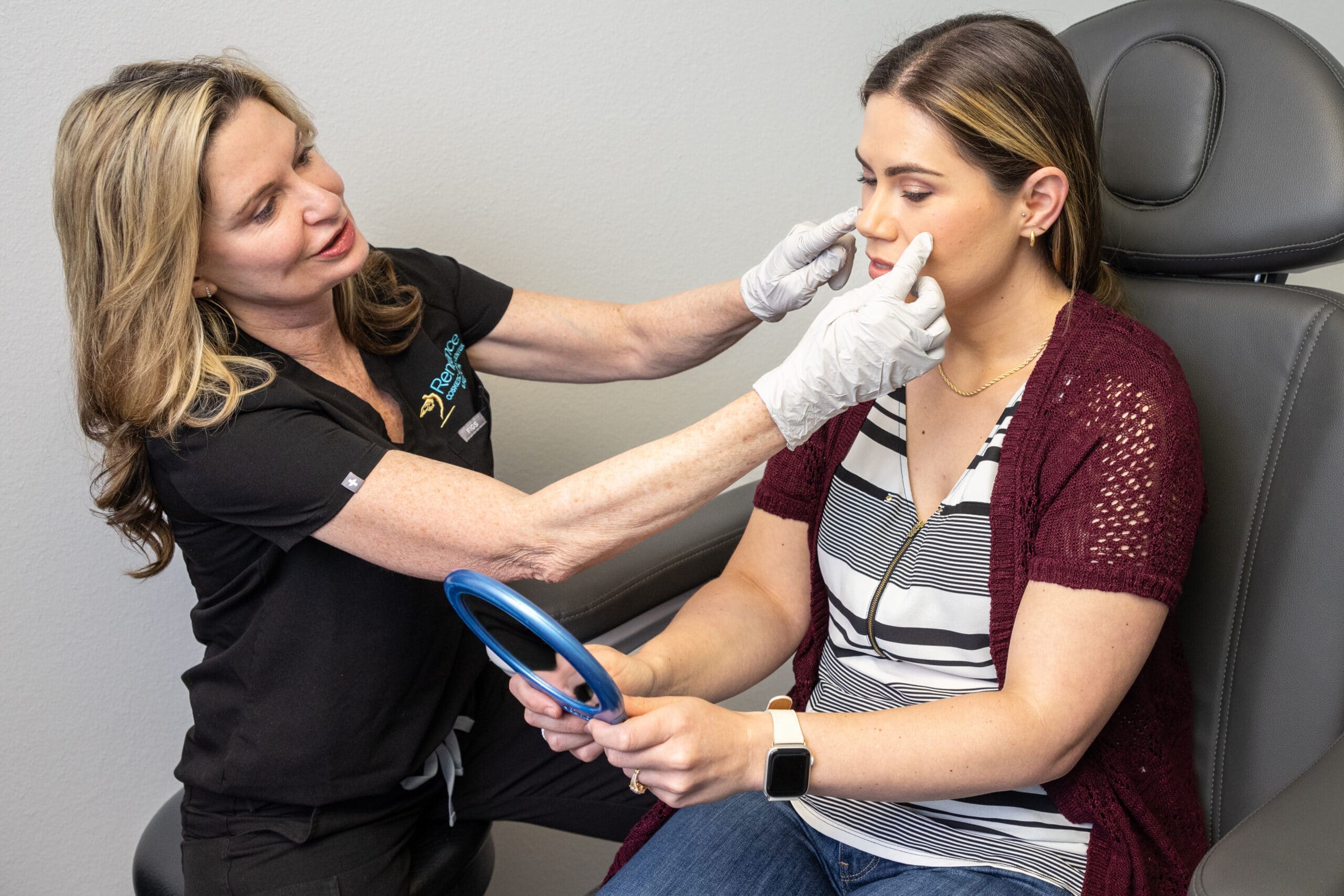 A medical professional wearing gloves examines a woman's face as she holds a mirror while sitting in a chair. The woman is wearing a striped shirt and a maroon cardigan. They are in a clinical setting.