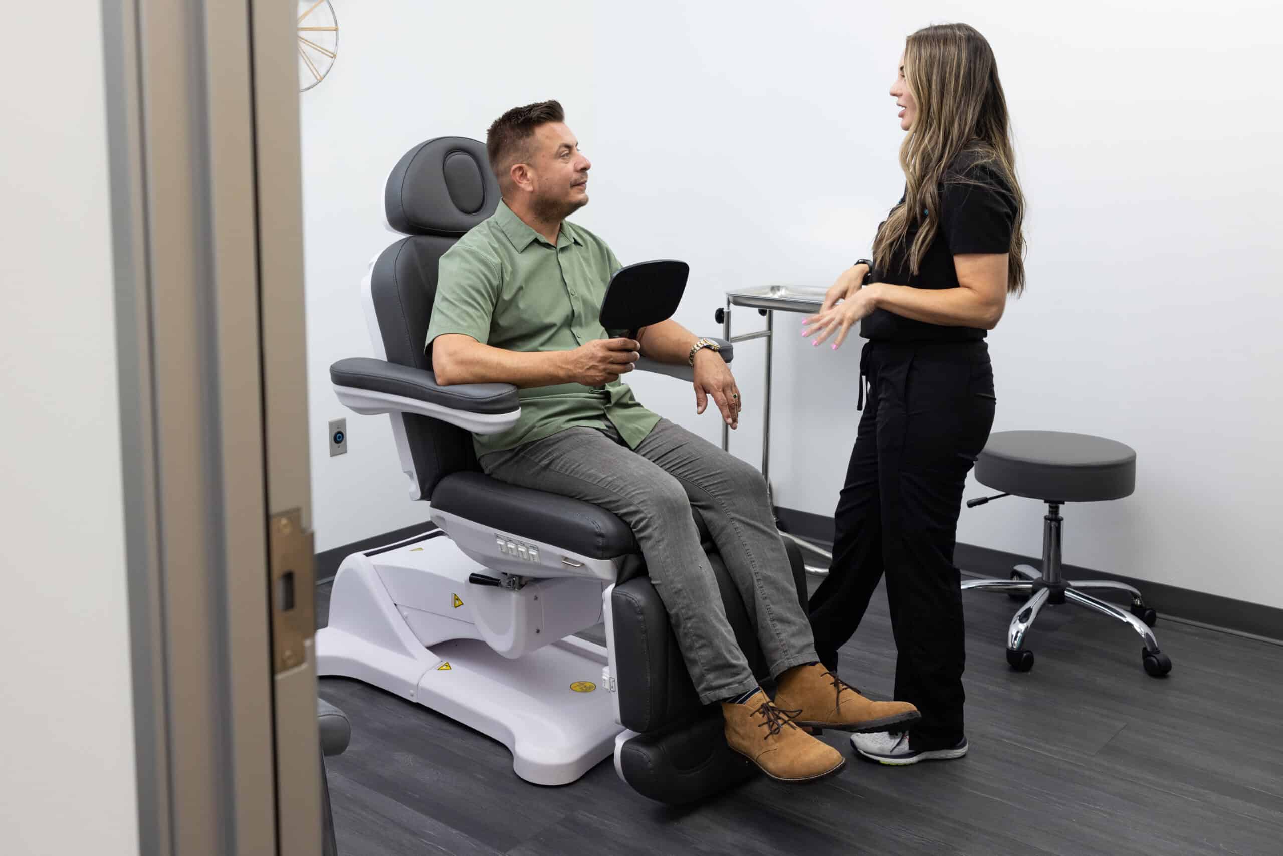 A man is seated in a medical examination chair holding a small mirror and looking at a female healthcare professional who is standing and talking to him. The room features medical equipment such as an adjustable stool and a small side table. The walls are white, and the floor is dark-colored wood.
