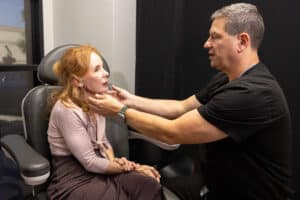 A healthcare professional is examining an older woman's neck while she sits in a medical chair. The professional is gently holding her chin and neck, appearing focused and attentive. The setting looks like a medical office with medical equipment and an examination environment. Both individuals are engaged in the medical assessment.