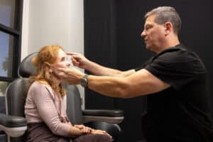 A man in a black shirt is examining the eyes of a woman with red hair seated in a chair. The woman is wearing a light pink top and looks towards the man. The setting appears to be a medical office.