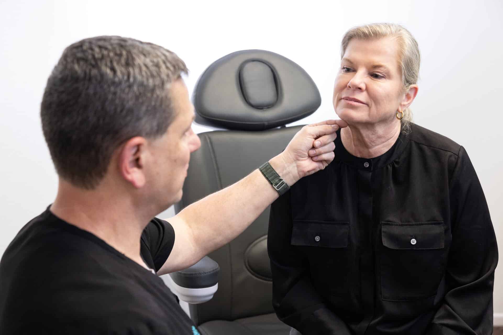 A man is examining a woman's neck in a clinical setting. The woman is seated on a chair and appears to be following the man's instructions while he gently touches her chin. Both are engaged in a medical assessment, with the man appearing to be a healthcare professional.