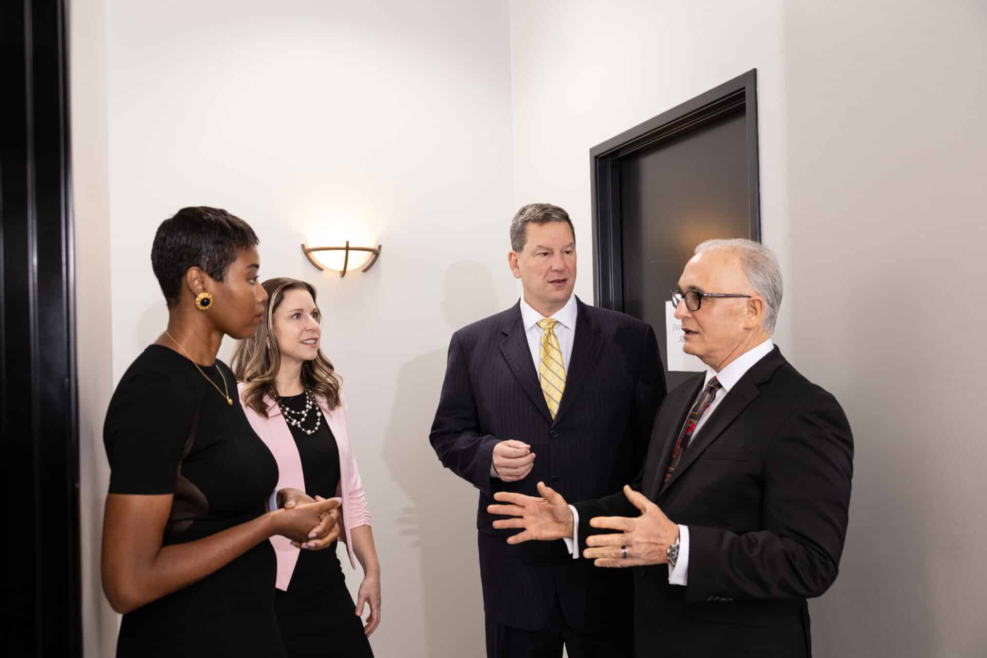Four professionally-dressed individuals are seen engaged in conversation in an office hallway setting. Three of them are standing together, listening attentively, while the man on the right speaks, making hand gestures. The lighting is bright and there is a wall sconce visible in the background.