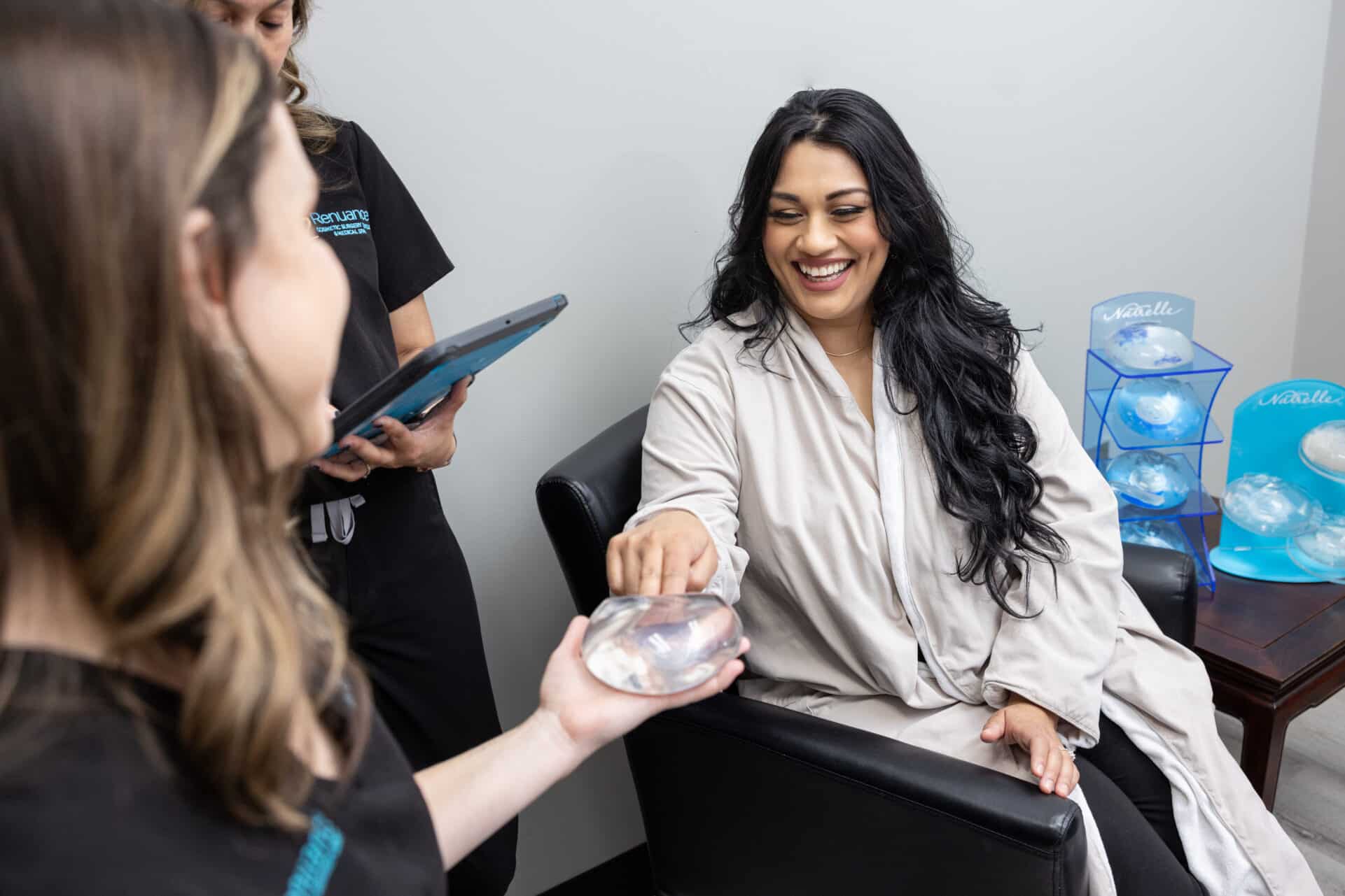 A woman with long dark hair is seated in a black chair, smiling and touching a breast implant that is being held by a medical professional wearing black scrubs. Another medical professional is standing nearby holding a tablet. On a table behind them, there are several more breast implants displayed. The setting appears to be a medical or consultation room.