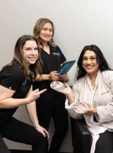 Three women are present in a medical office setting. One woman is seated and holding two clear, spherical objects. She is smiling and dressed in a white lab coat. The two other women, standing on either side of her, are wearing black uniforms and also smiling. One of them is pointing towards the seated woman, while the other holds a tablet. The background is a plain, light-colored wall.
