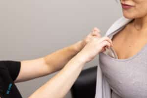 A person is being examined by a healthcare professional. The professional is using one hand to palpate the patient's left upper chest area over their clothing while the other hand rests on the patient's arm. The patient is wearing a light gray sleeveless top.