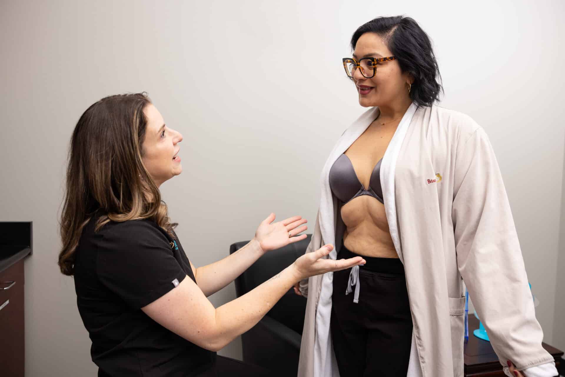 A woman in a black outfit with long hair is sitting and talking to another woman who is standing. The standing woman is wearing glasses, a light-colored robe, and a bra, revealing her midsection. They appear to be in a medical or consultation setting and engaging in a conversation.