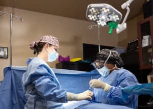 Two medical professionals in surgical scrubs and masks performing a surgery under bright surgical lights in an operating room. The room is equipped with medical supplies and devices.