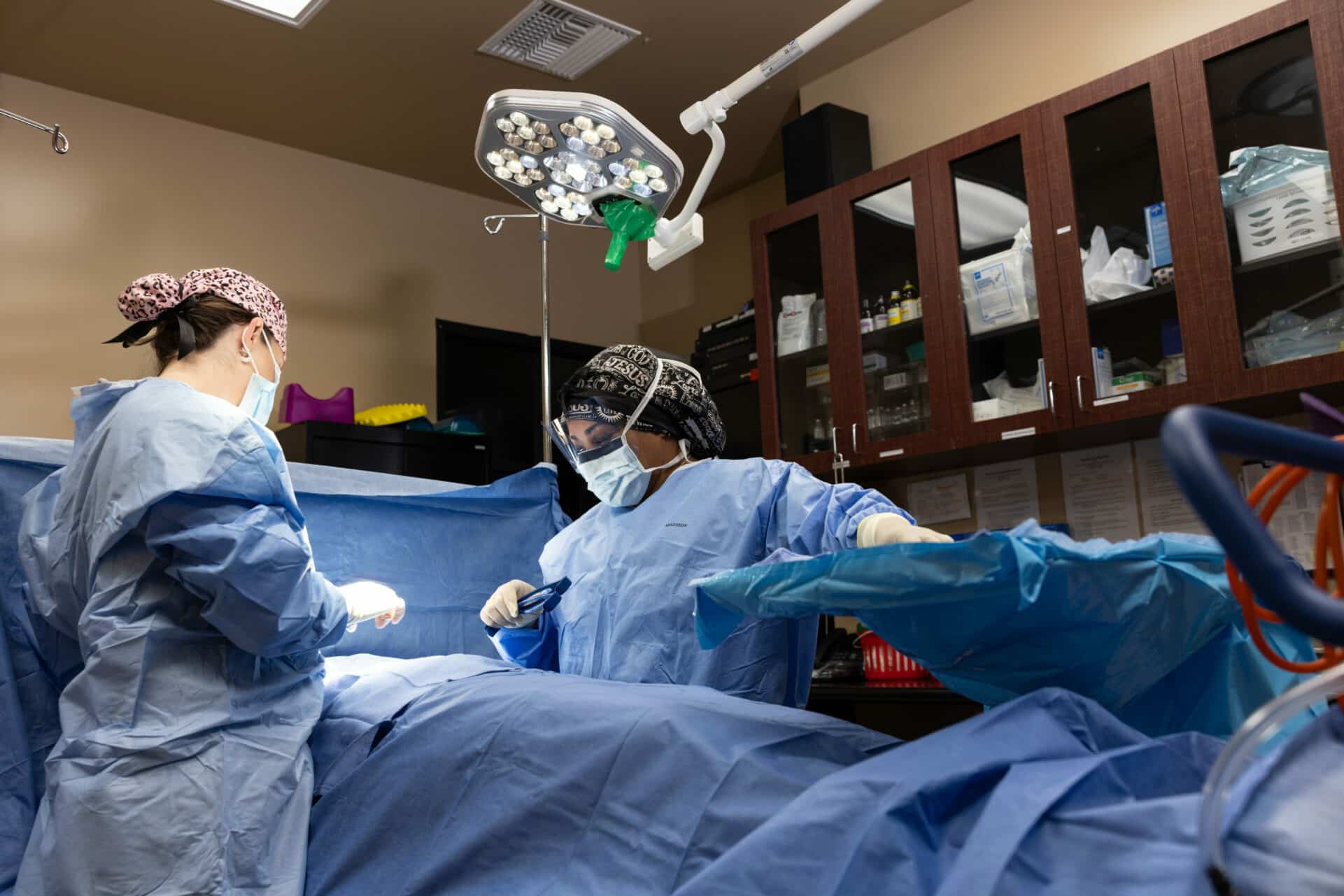 Two medical professionals in surgical attire perform a procedure on a patient in an operating room. Both are wearing masks and surgical caps. One of them appears to be making an incision while the other assists by holding tools. The room is equipped with surgical lights and various medical equipment and supplies are visible in the background.