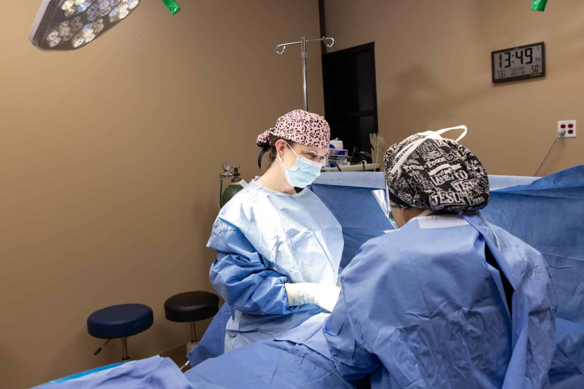 Two healthcare professionals, dressed in surgical scrubs and masks, are performing a medical procedure in a well-lit operating room. One person is standing and focusing on the task, while the other is seated or leaning over. Surgical equipment and a clock on the wall displaying the time 13:49 are visible in the background.