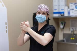 A medical professional in black scrubs, a pink leopard print cap, and a blue surgical mask is standing in a clinical setting with hands clasped together. The background includes medical supplies and equipment.