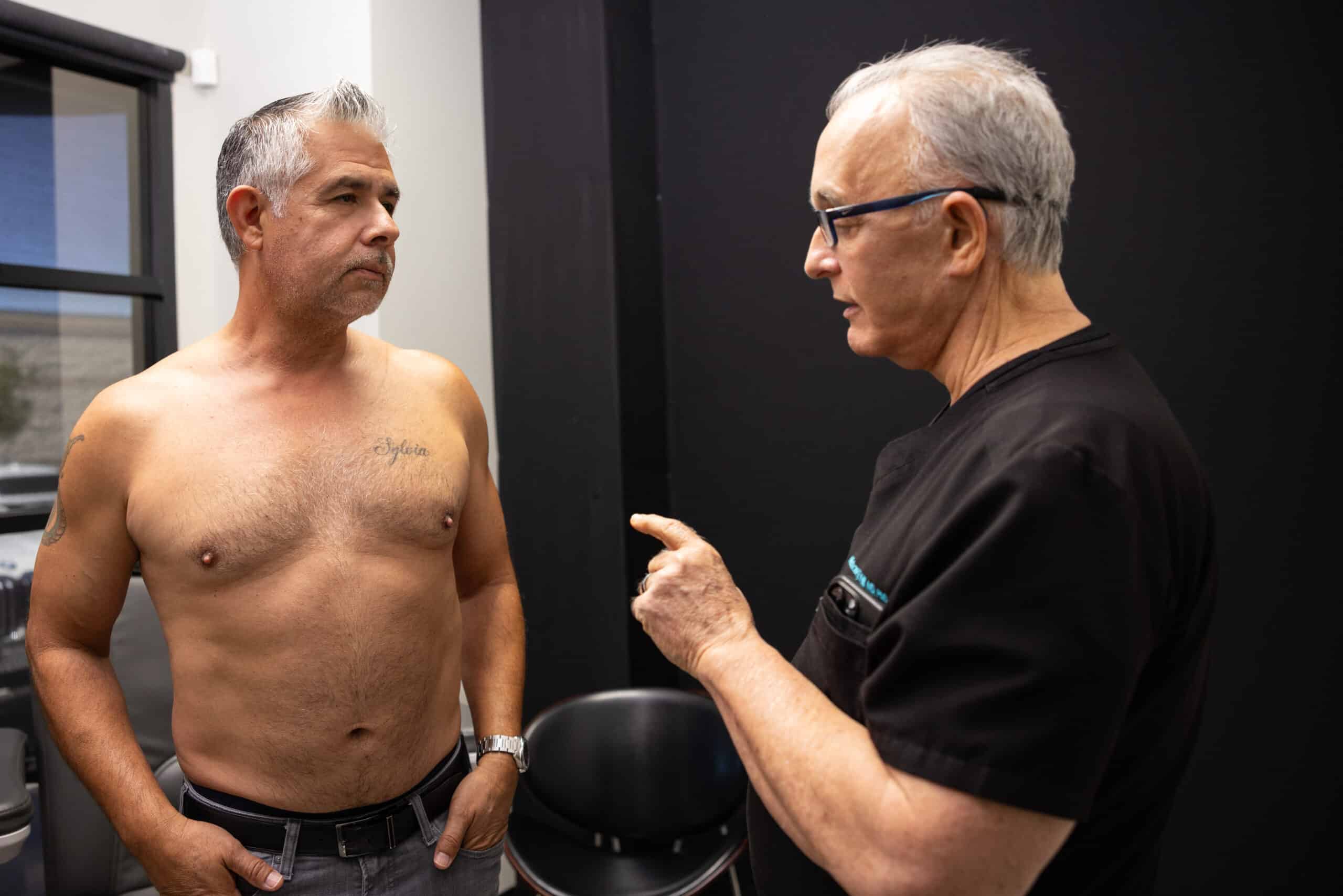 A shirtless man with gray hair stands facing a man wearing glasses and black medical scrubs. The man in scrubs is gesturing with his hand and seems to be explaining something to the shirtless man. They are in a room with dark walls and a window in the background.