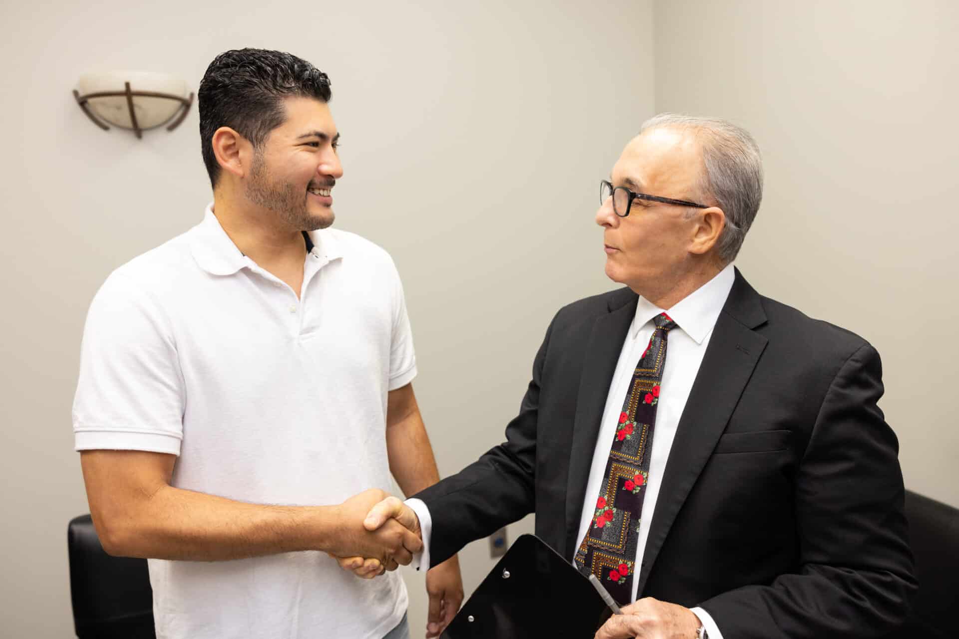 A younger man wearing a white polo shirt is shaking hands with an older man in a suit and tie. The older man is holding a black folder under his left arm. They are standing in a room with beige walls, light fixture on the wall, and sitting area furniture in the background. Both men are smiling.