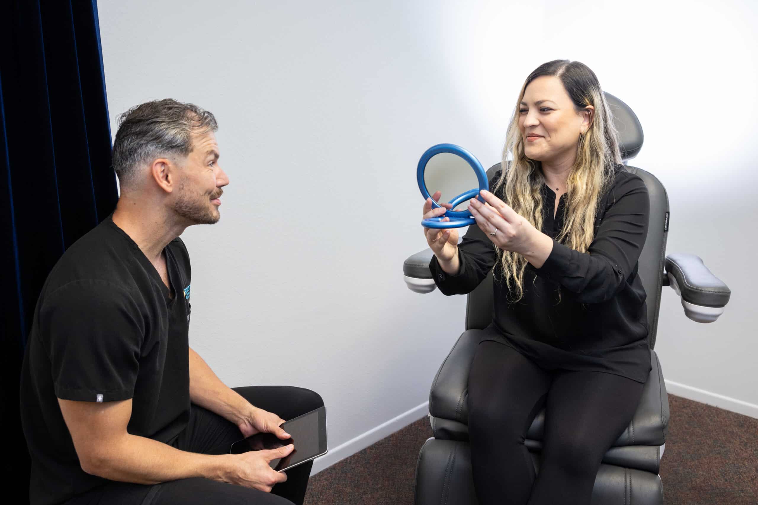 A woman with long blonde hair sits in a chair holding a round mirror and looking at it. A man with short, graying hair holds a tablet and sits nearby, engaging in conversation with the woman. Both are dressed in black clothing and are in a clinical or office setting.