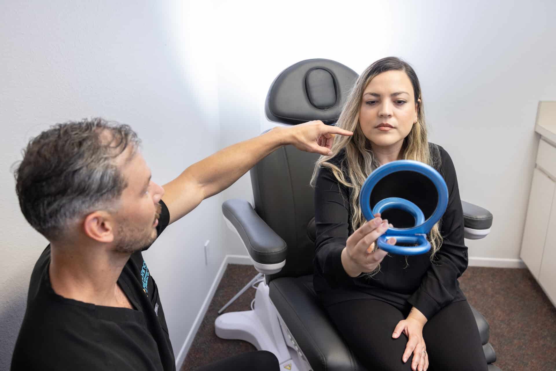 A woman sits in a chair holding a handheld mirror. A man, seated next to her, is pointing towards her face as if explaining or discussing something. They appear to be in a medical or professional office environment.