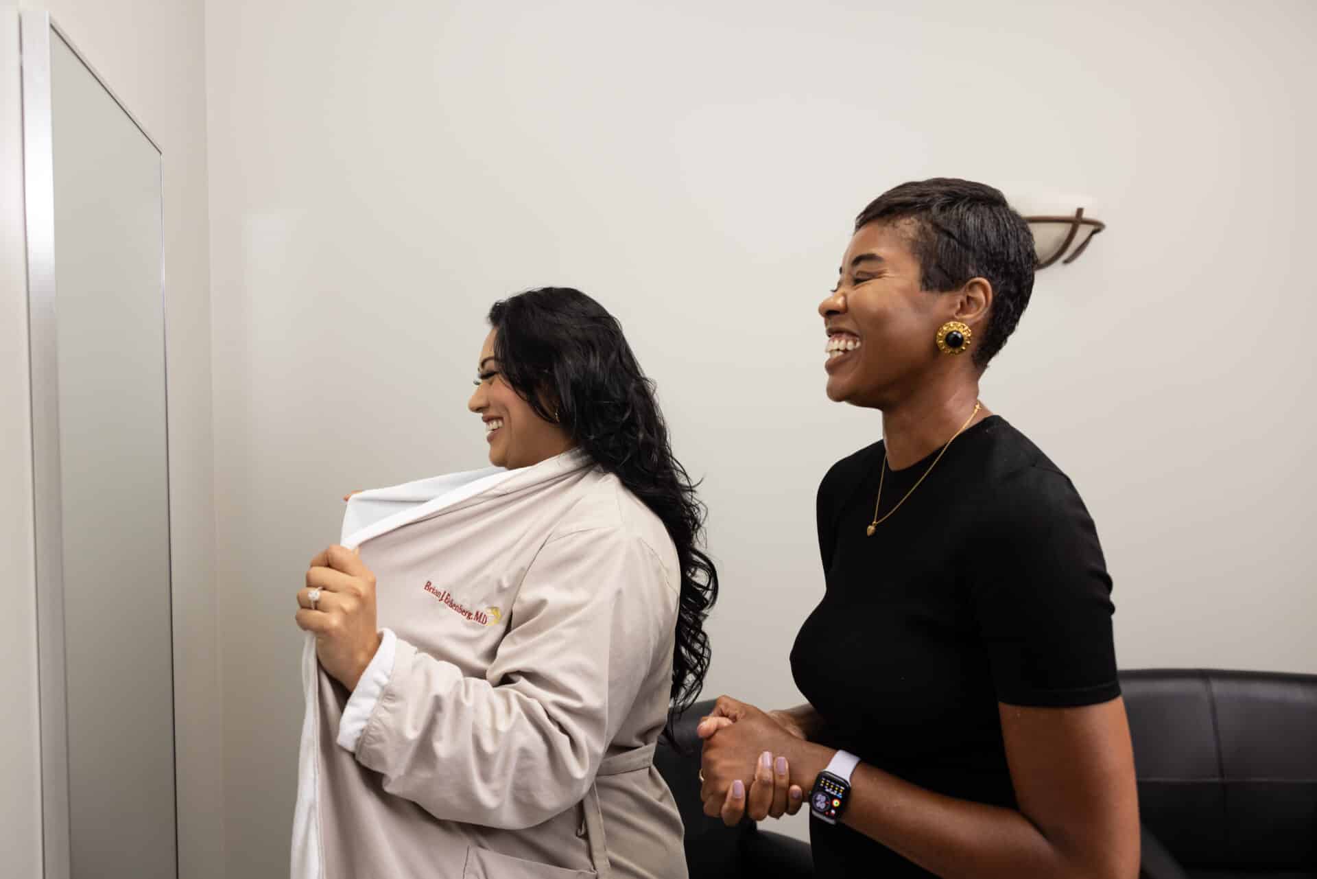 Two women stand indoors, smiling and laughing. One woman on the left is wearing a beige robe and appears to be adjusting or examining it. The other woman on the right, dressed in a black shirt and wearing an Apple Watch, is looking towards her with a joyful expression. The background is plain with neutral tones.