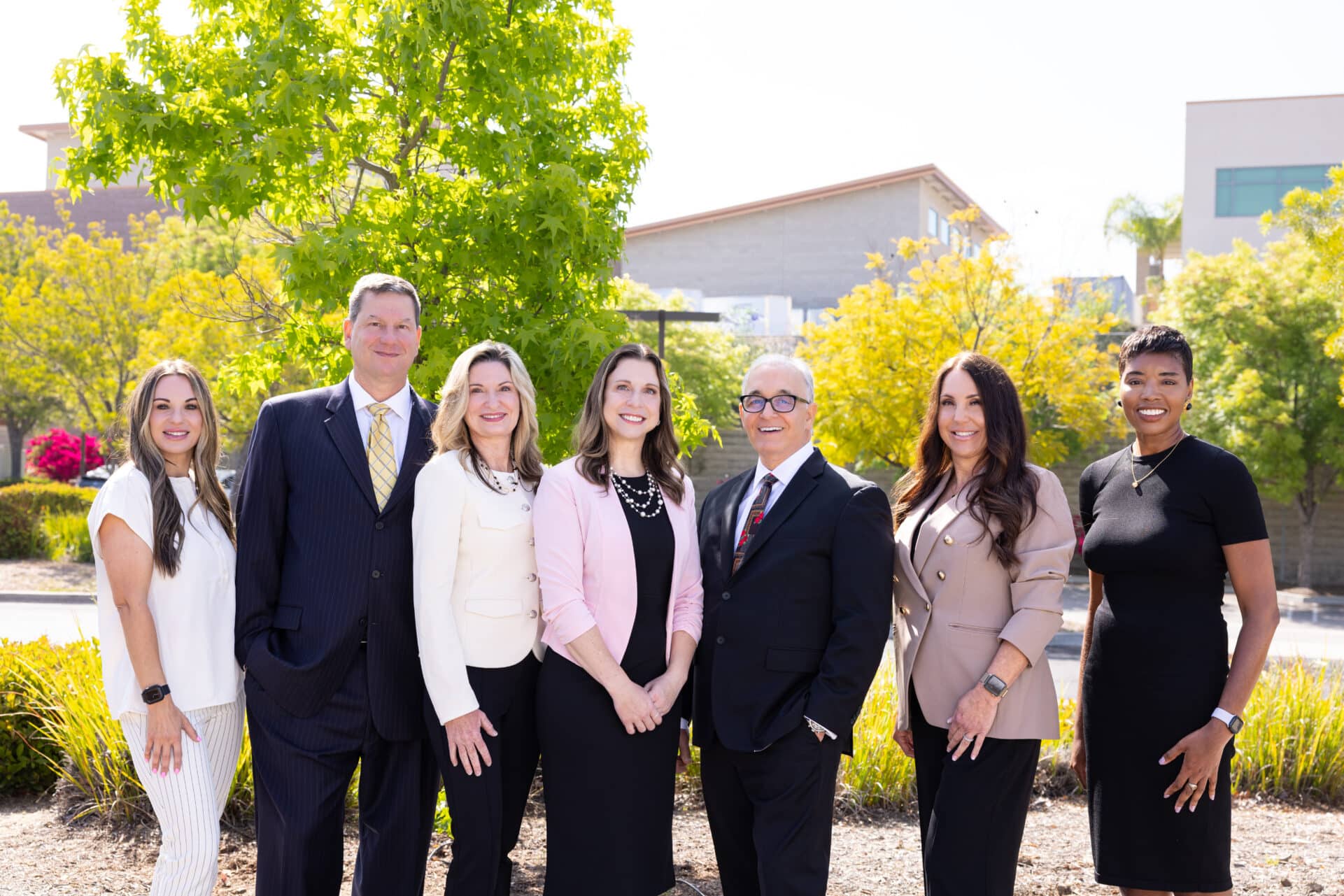 A group of seven professionally dressed people is standing outdoors, smiling and posing for the camera. They are positioned in a semi-circle in front of greenery and trees, with some buildings visible in the background. The group includes both men and women, dressed in suits, blouses, and dresses, suggesting a formal or corporate setting. The sky is clear and the lighting is bright, indicating it is a sunny day.