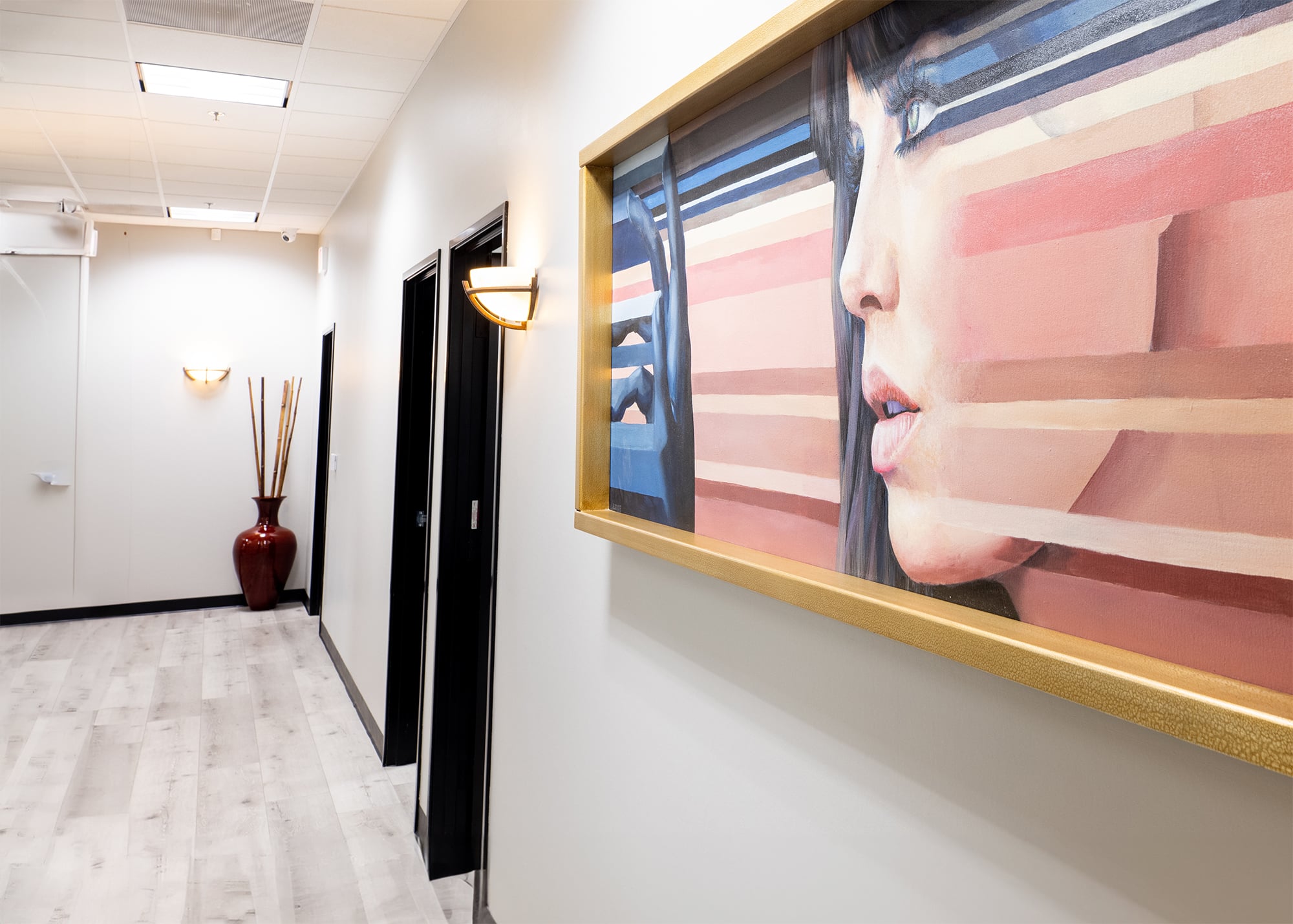 A hallway with light-colored wooden flooring and white walls. The hallway has closed black doors lining one side and a large contemporary painting of a woman's face with abstract geometric lines on the opposite wall. There is a modern wall sconce light fixture under the painting. At the end of the hallway, a tall red vase with decorative branches is positioned on the floor. The ceiling is outfitted with recessed lighting.