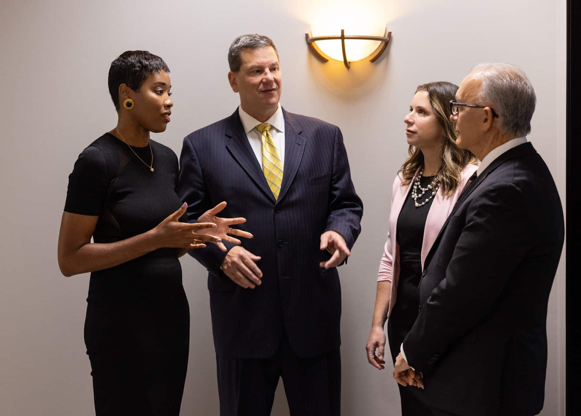 A group of four people standing and having a discussion. They are dressed formally, with three of them wearing suits and one person in a dress. They are inside a room with beige walls, and a wall light fixture is visible in the background.