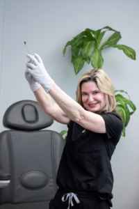 A woman wearing a black outfit and white gloves is holding a syringe up in a bright, clinical-looking room. She has blonde hair and is smiling at the camera. In the background, there is an empty gray chair and a large green plant.