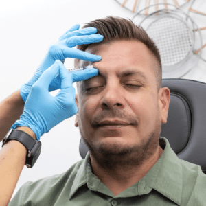 A man receiving a cosmetic injection on his forehead from a medical professional wearing blue gloves. The man is sitting in a chair with his eyes closed and a relaxed expression. The background features a modern wall decoration, partially visible.
