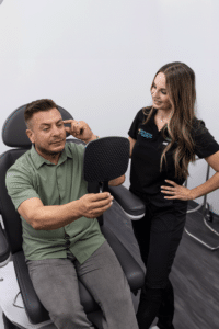 A man sits in a chair, holding a handheld mirror and touching his face, while a woman stands beside him, smiling. The woman is wearing a black uniform with a company logo, while the man is dressed casually in a green shirt and grey pants. The setting appears to be a clinic or medical office, with white walls and medical equipment in the background.