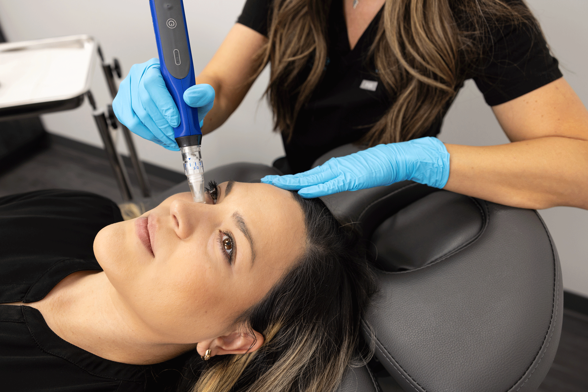 A woman is lying back in a reclining chair during a skincare procedure. A professional wearing black clothing and blue gloves is using a microneedling device on the woman's forehead. The woman has her eyes closed and appears relaxed. In the background, a white tray with tools can be seen.