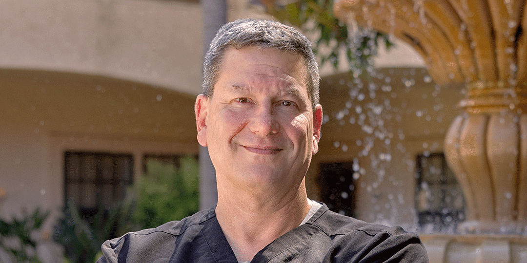 A man with short hair is smiling and standing outdoors in front of a fountain. He is wearing black scrubs and has his arms crossed. The background features greenery and a building.