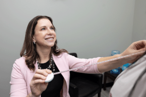 A woman is smiling and holding a measuring tape around another person's body. She appears to be in a professional office setting, sitting on a chair with a dark fabric. The woman has long brown hair and is wearing a light pink blazer over a black top, accessorized with a long necklace. The background features office furniture, including a chair and some items on a table.