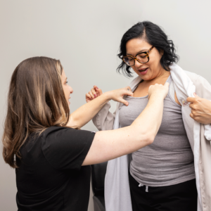 A woman is sitting down, wearing a light-colored tank top and a white coat over her shoulders. Another woman, standing to the left, wearing a black top, is assisting her. The standing woman appears to be adjusting or examining the tank top on the seated woman. The background is plain and light-colored.