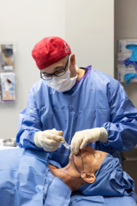A surgeon in blue scrubs and a red cap is performing a procedure on a patient, also dressed in blue surgical attire, who is lying on an operating table. The surgeon is holding a surgical instrument and appears to be focused on the task. The room is equipped with medical supplies and equipment.
