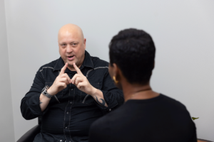 A bald man wearing a black shirt is sitting and gesturing with his hands as he talks to a person with short hair, seen from behind. The interaction appears to be in a professional or office setting with a white wall in the background.
