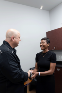 A man and a woman are shaking hands in an office setting. The man has a bald head and is wearing a dark jacket, while the woman has short hair and is dressed in a black, short-sleeved top. Both are smiling as they interact. The background reveals office furniture such as cabinets and a desk.