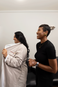 Two women are standing indoors, smiling and laughing. The woman on the left is wearing a light-colored coat with text on it and has long, dark hair. She appears to be putting the coat on or adjusting it. The woman on the right has short, dark hair and is dressed in a black outfit, accessorized with a watch and gold earrings. Both are engaging in a cheerful conversation in what seems to be a comfortable setting.