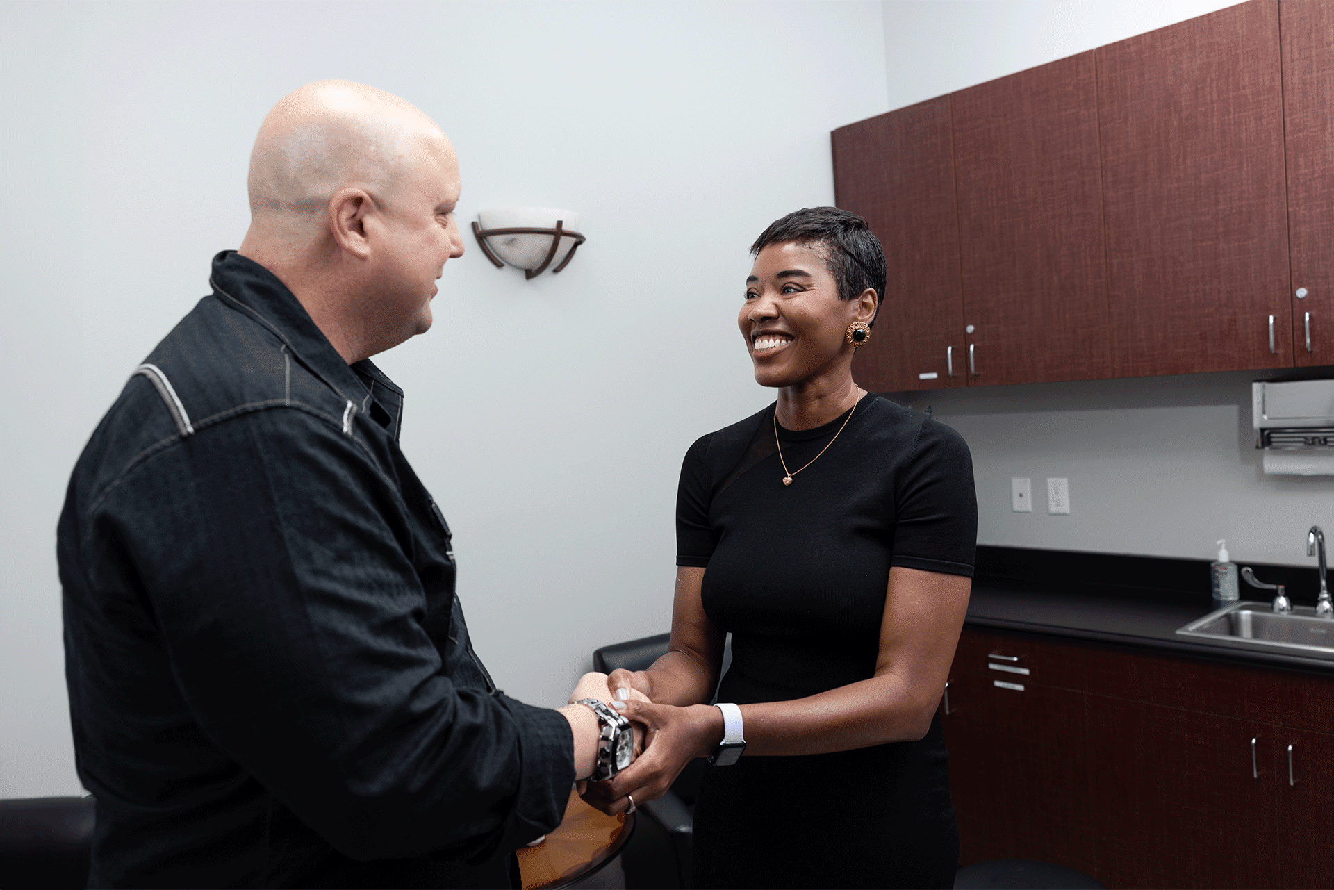 A man and a woman shaking hands in an office setting. The woman, dressed in a black top, is smiling warmly, while the man, wearing a dark jacket, also appears to be smiling. The office has minimalist decor, with a few cabinets, a small sink, and a wall-mounted light.