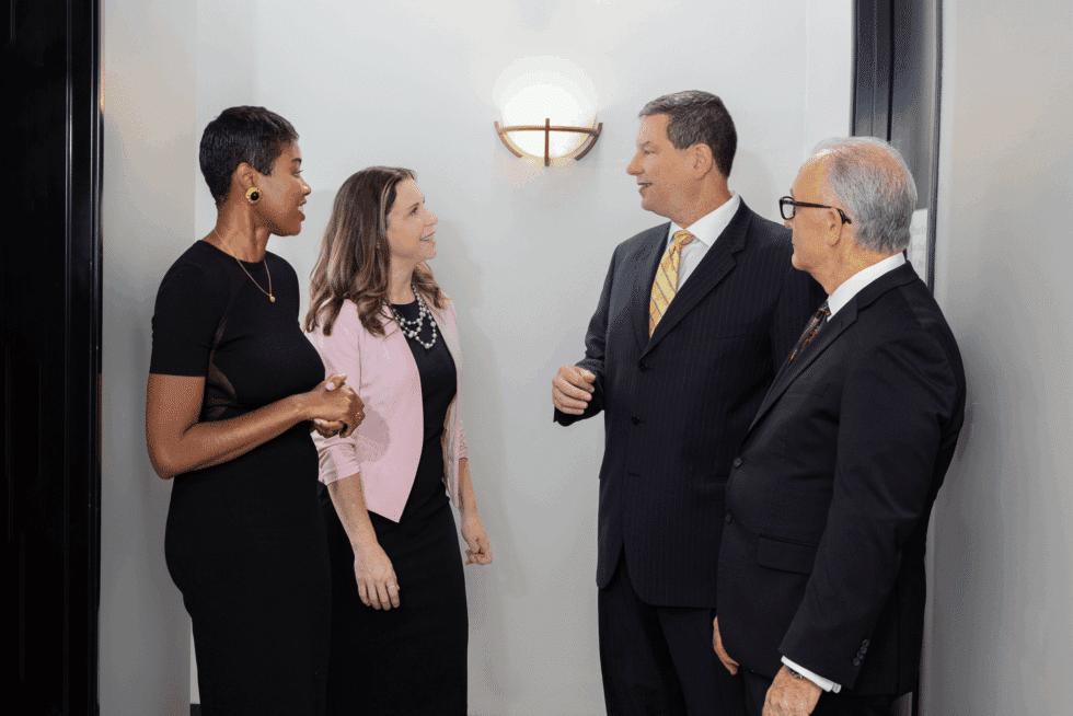 Four individuals are engaged in a conversation in a hallway. From left to right, the first individual is wearing a black dress, the second is in a pink jacket over a black dress, the third is in a dark suit with a yellow tie, and the fourth is in a dark suit and tie. They are all smiling and appear to be having a pleasant discussion. The hallway has a light fixture illuminating the area.