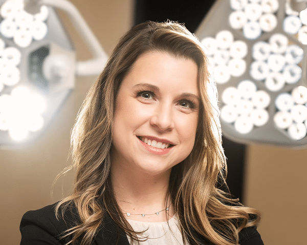 A woman with long, wavy, blonde hair is smiling and looking at the camera. She is wearing a black blazer over a light-colored shirt and a delicate necklace. The background consists of bright lights with circular, flower-like arrangements, suggesting a professional or medical setting.